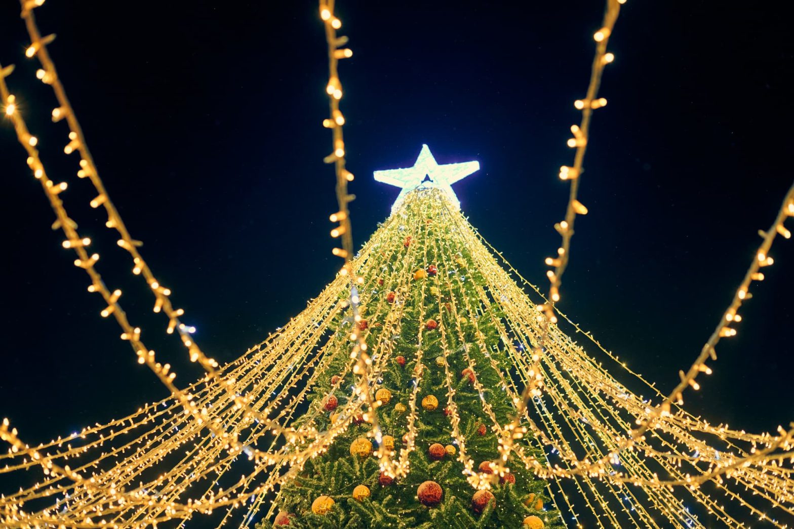 Ένα ψηλό, λαμπερό χριστουγεννιάτικο δέντρο στολισμένο με χρυσά φωτάκια και πολύχρωμες μπάλες.