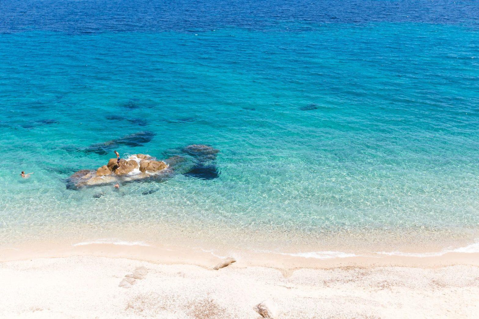 bright blue water and light colored sand with people standing on a rock at the beach