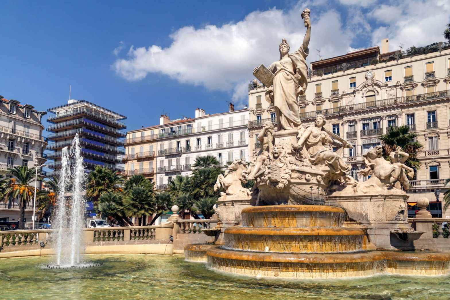 Fountain with sculptures and cascading water in a sunny square with palm trees and traditional European buildings in the background.