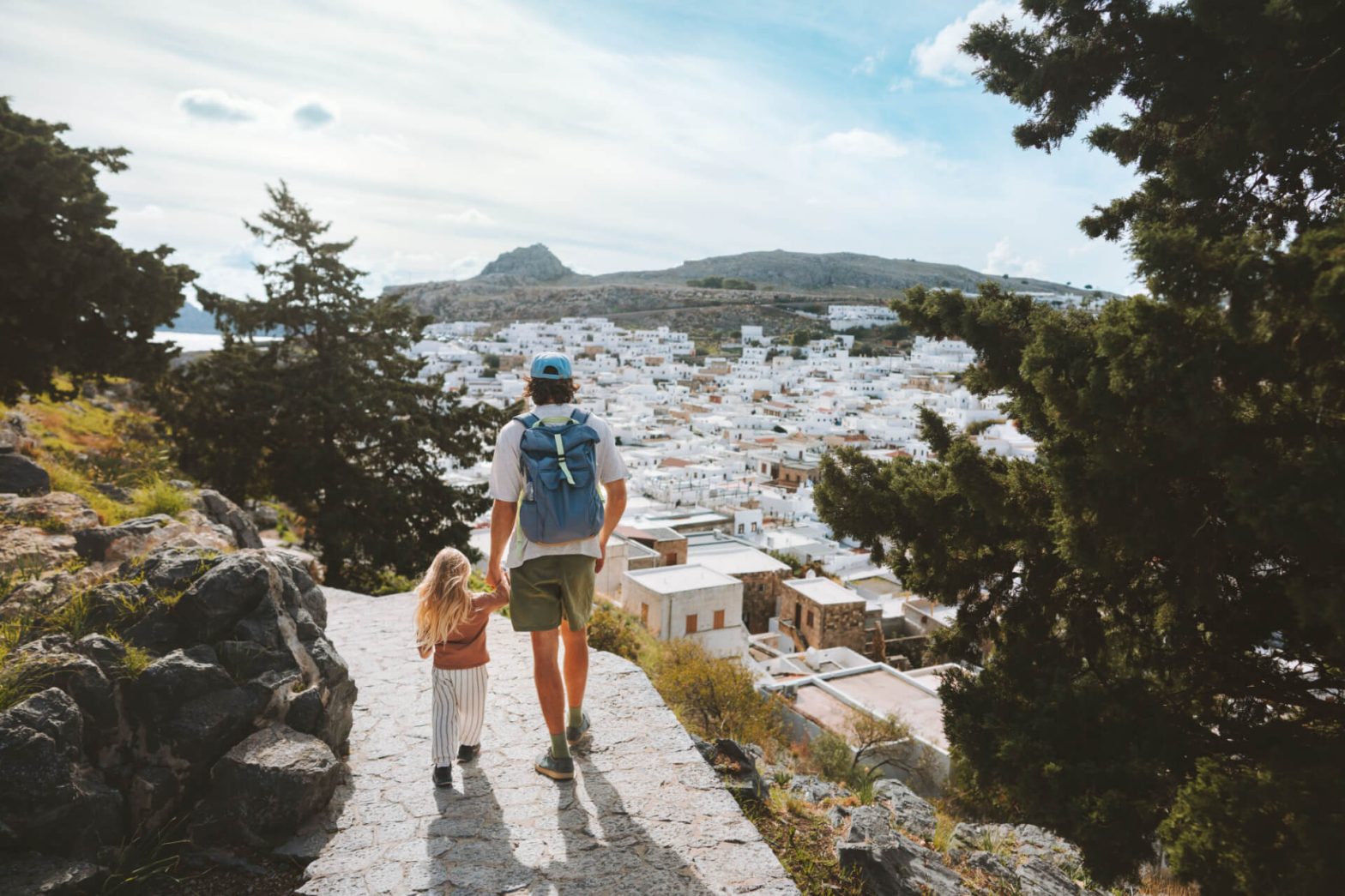 An adult and child walk hand in hand down a stone path overlooking a sunlit white village amidst greenery on a Greek island.