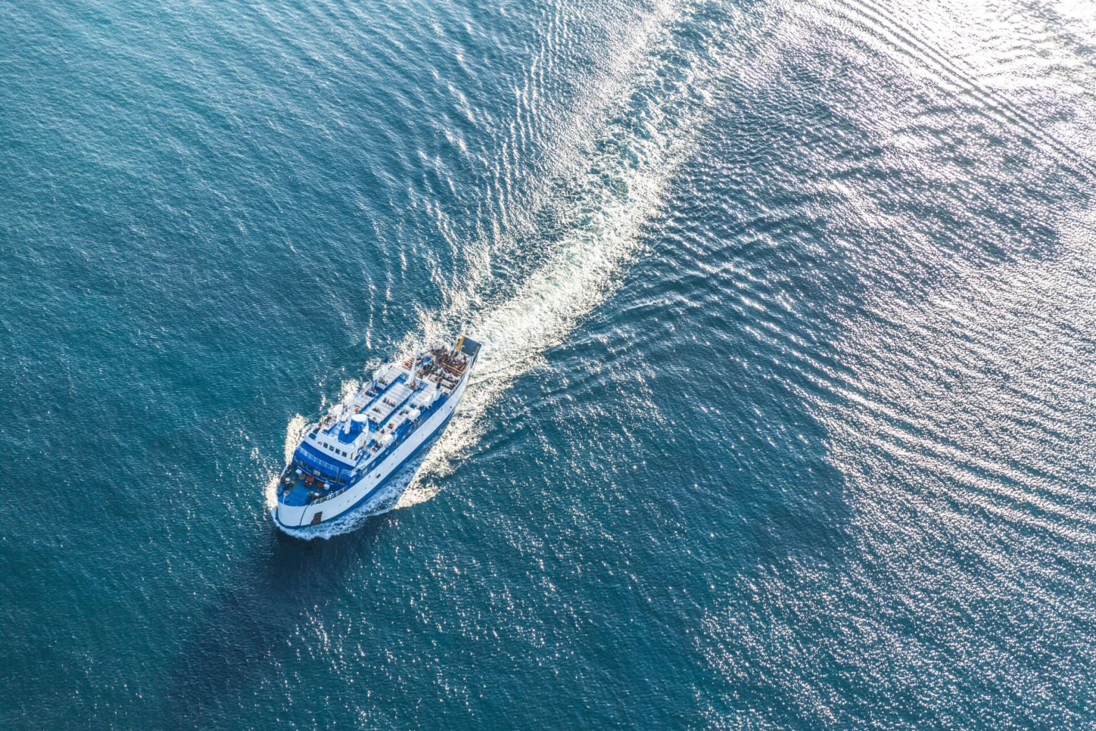 A ferry cutting through the blue waters of the Aegean Sea, leaving a frothy wake behind, captured from an aerial perspective in bright daylight.