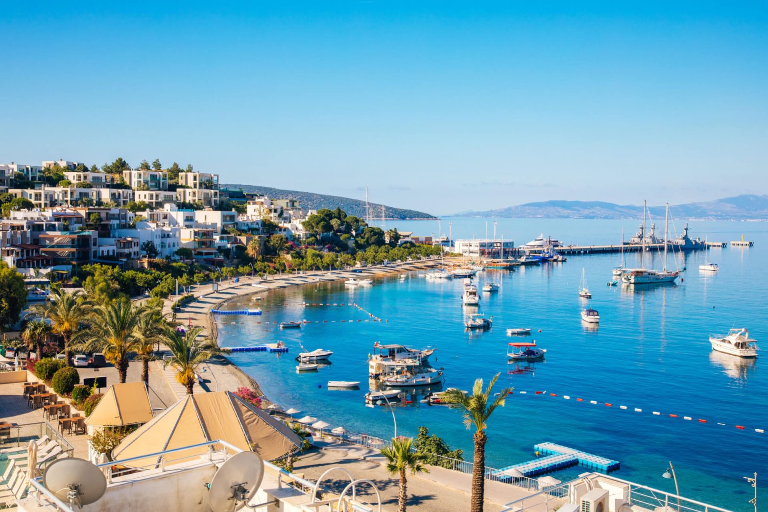 A serene bay with boats, a sandy shore, and modern hillside houses with a backdrop of clear skies and distant mountains.