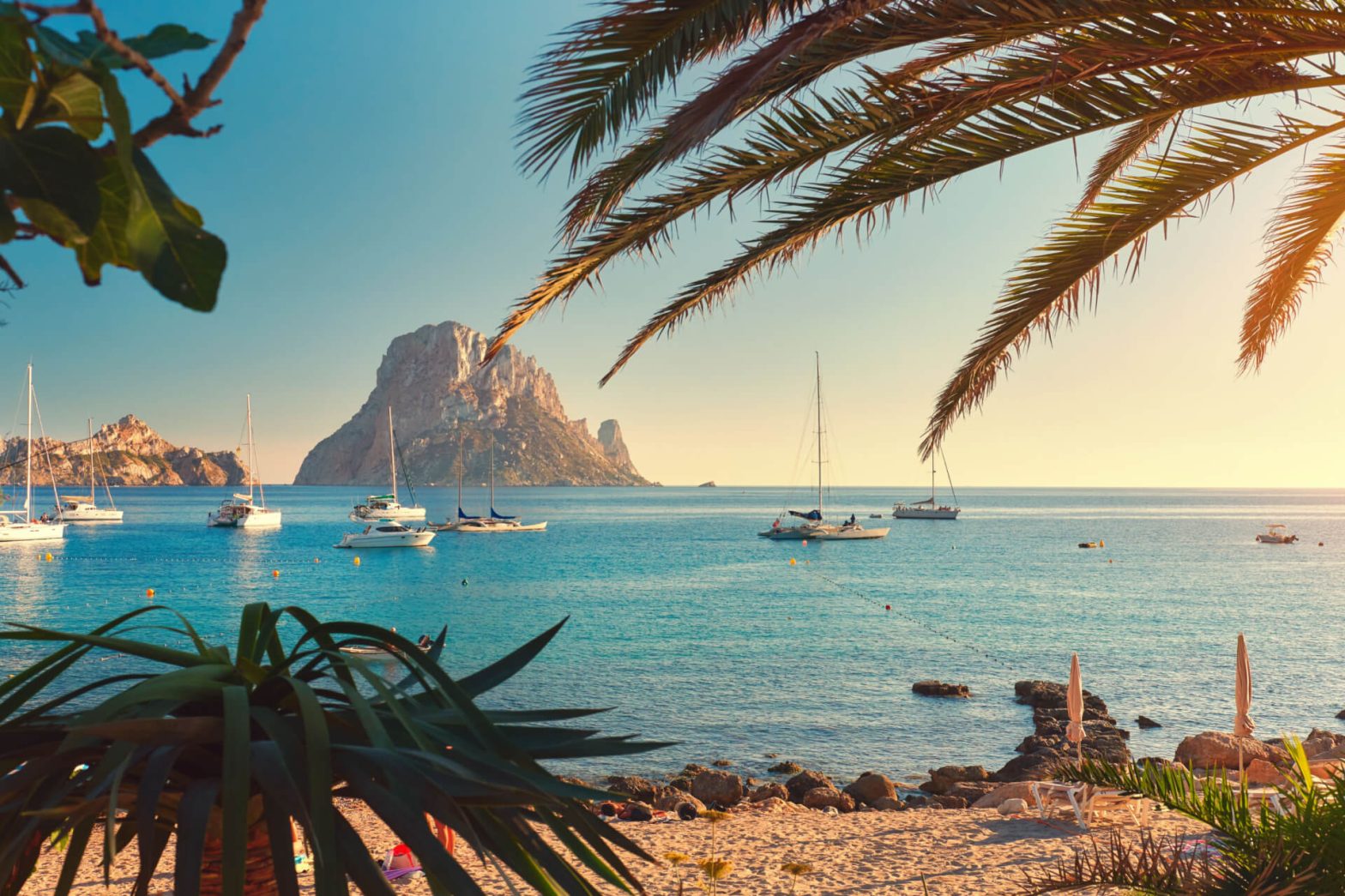 A tranquil Ibiza beach with palm fronds in the foreground and yachts floating near the mystical rock formation of Es Vedra under a clear blue sky.