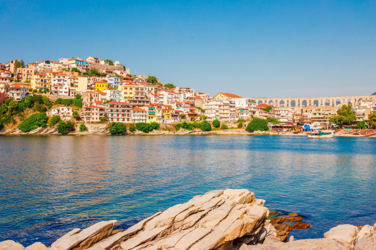 A photo of a harbor town with colorful houses on a hillside overlooking a blue Aegean Sea.