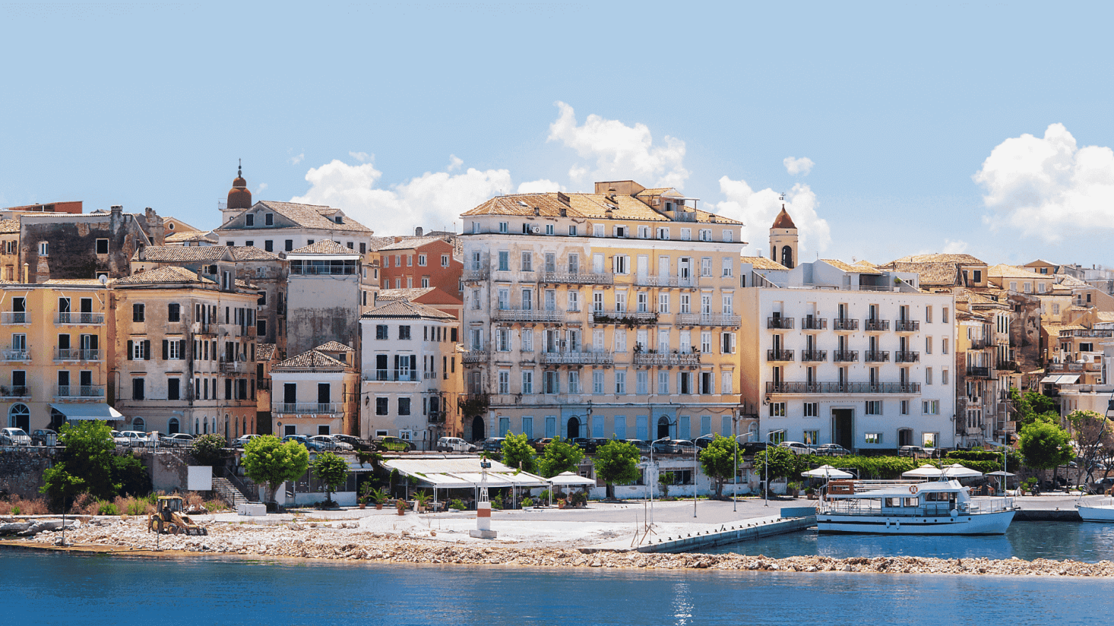 κτηρια σε διαφορα χρωματα σε σημειο της κερκυρας διπλα απο την θαλασσα οπου στον πεζοδρομο περπαταει κοσμος