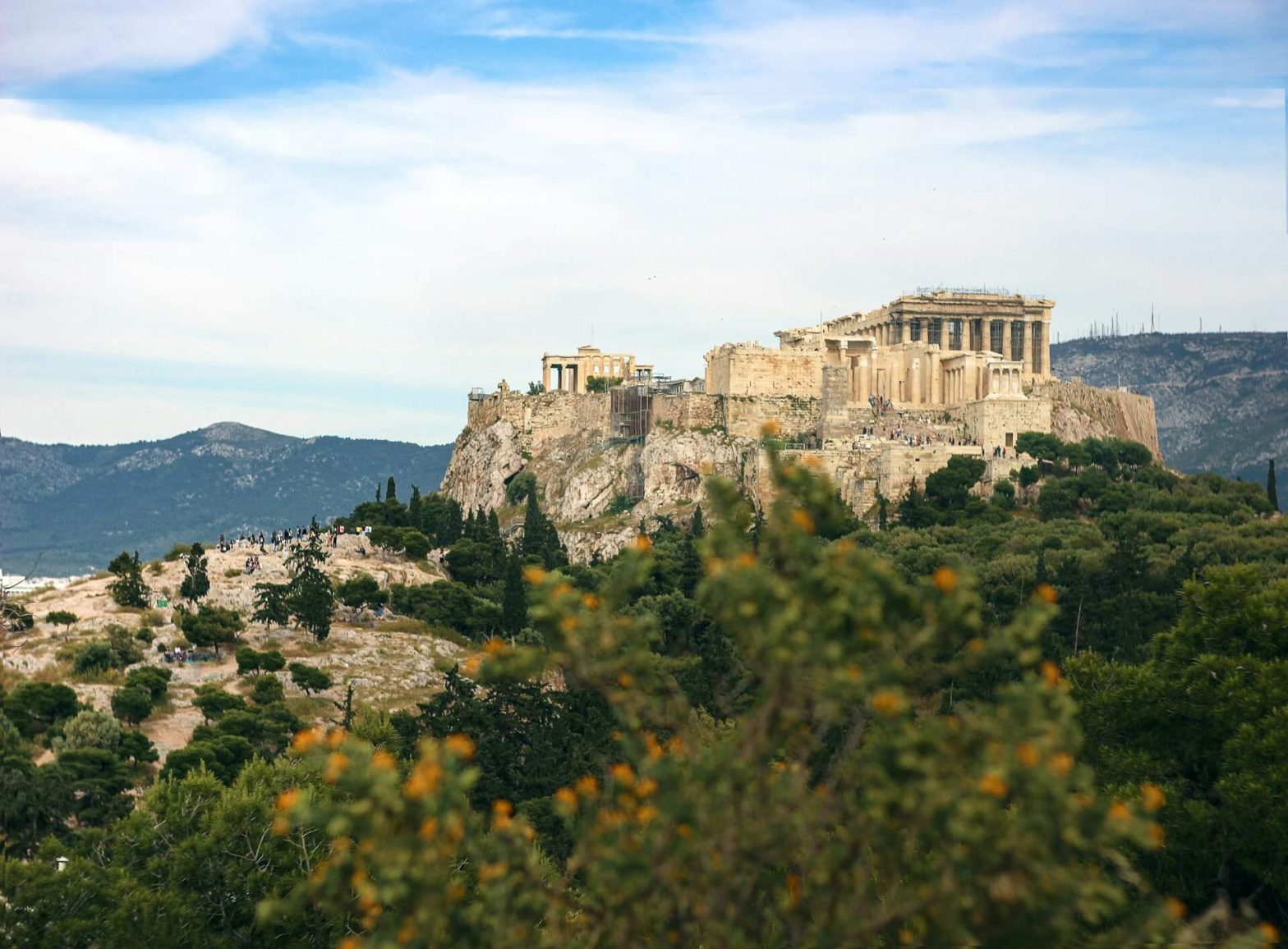 acropolis of athens, with green bushes in the foreground and wispy white clouds in the sky
