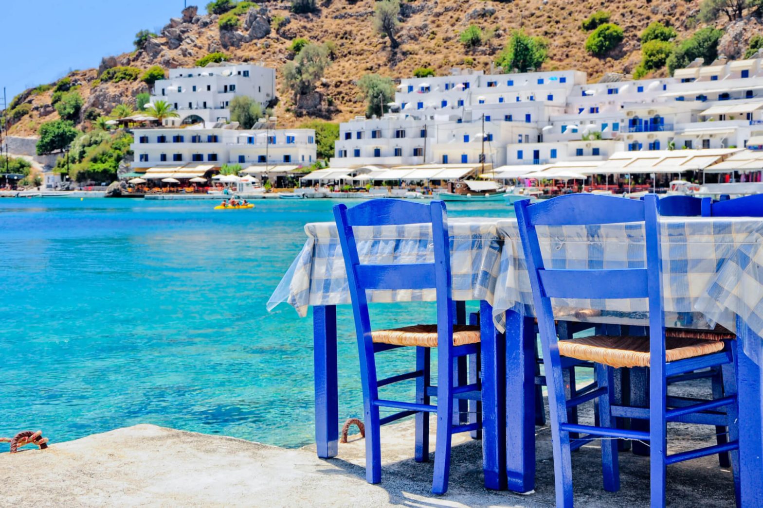 Seaside Greek café with blue chairs, checkered tablecloths, overlooking a bay with white buildings on a hill and boats on azure waters.
