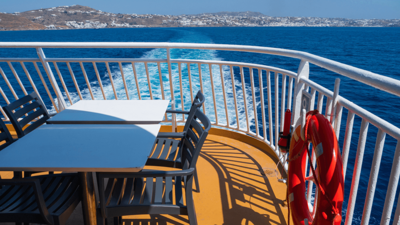 γκρι καρεκλες με λευκο τραπεζακι στην μεση στο καταστρωμα του πλοιου ενω στο βαθος μπορεις να δεις το μπλε απο τη θαλασσα καθως το πλοιο χαραζει τα κυματα