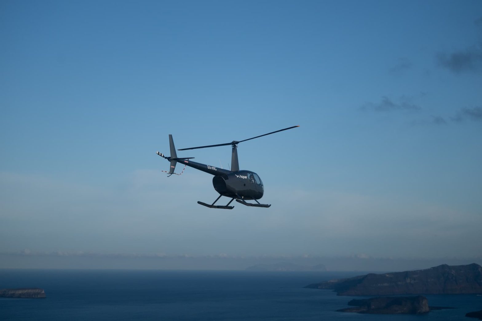 A black helicopter flies over the dark blue sea with a clear blue sky in the background.
