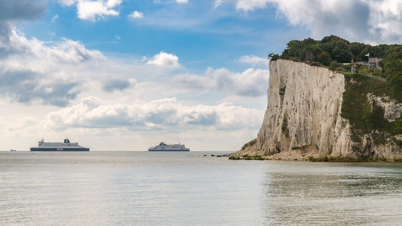 large white rock formation that is the cliffs of dover on the right side while two large ferry ships sit on the horizon sailing from france to the UK