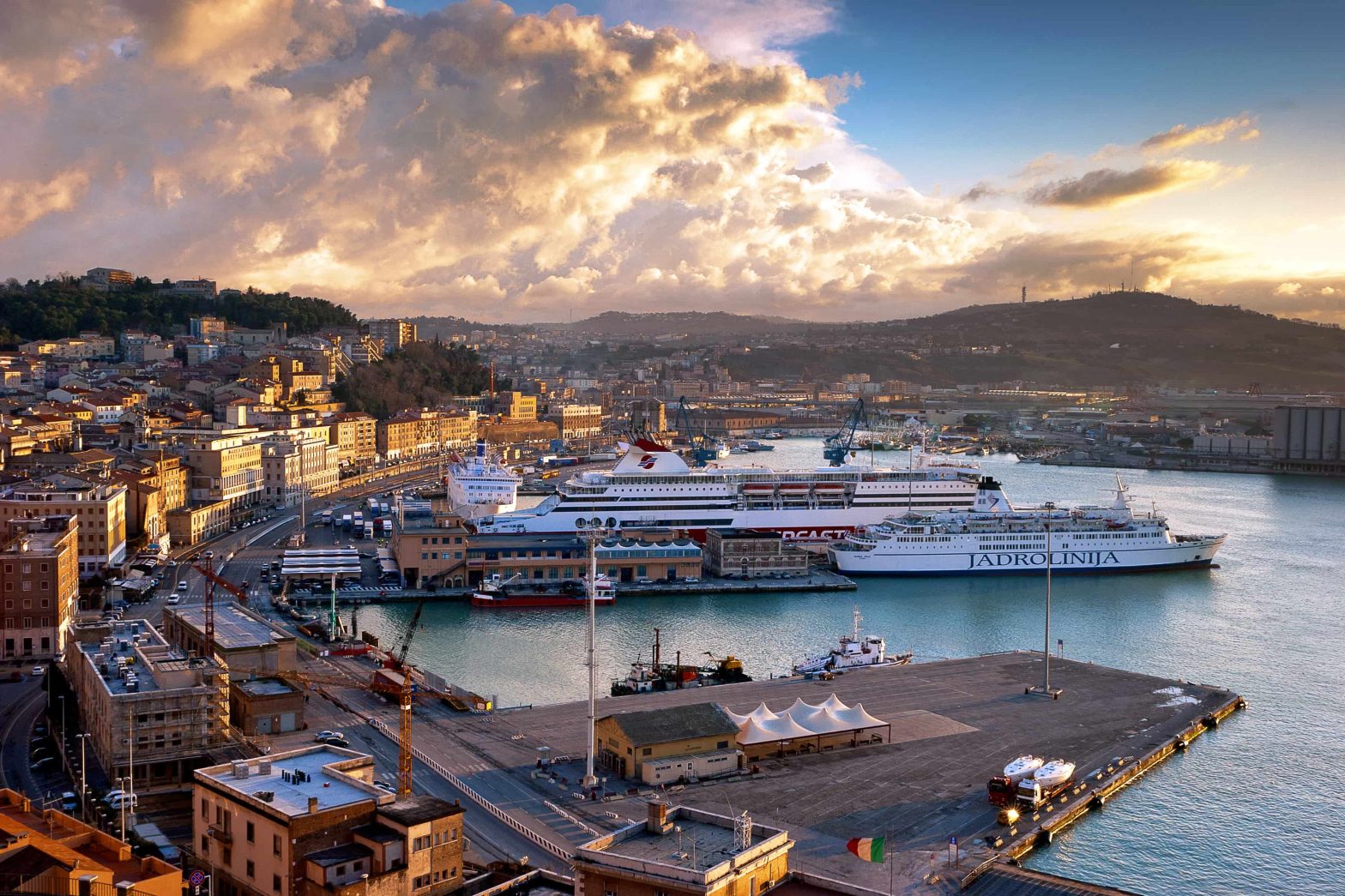 Aerial view of Ancona, Italy, featuring its bustling harbor, ferries, and a scenic hillside backdrop at sunset.