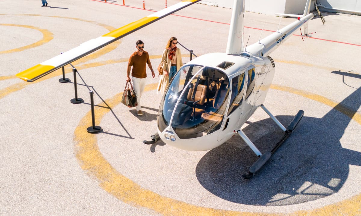 A stylish couple walks towards a white helicopter with yellow accents on a sunlit helipad. The luxury travel setting highlights Hoper's exclusive air transfer service.