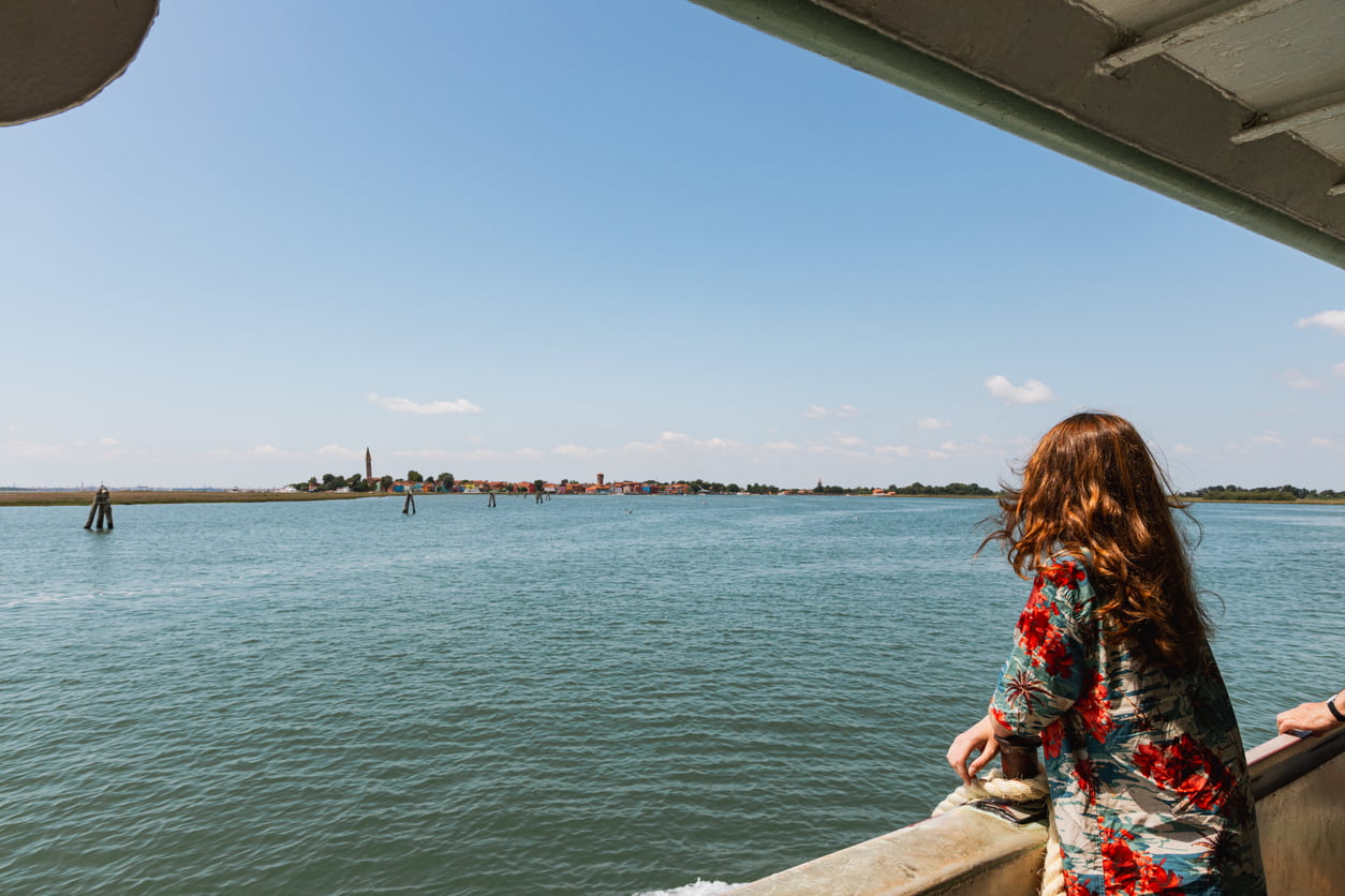 Γυναίκα απολαμβάνει τη θέα από το κατάστρωμα πλοίου στη λιμνοθάλασσα της Βενετίας, με φόντο ένα νησί και γαλήνια γαλάζια νερά.