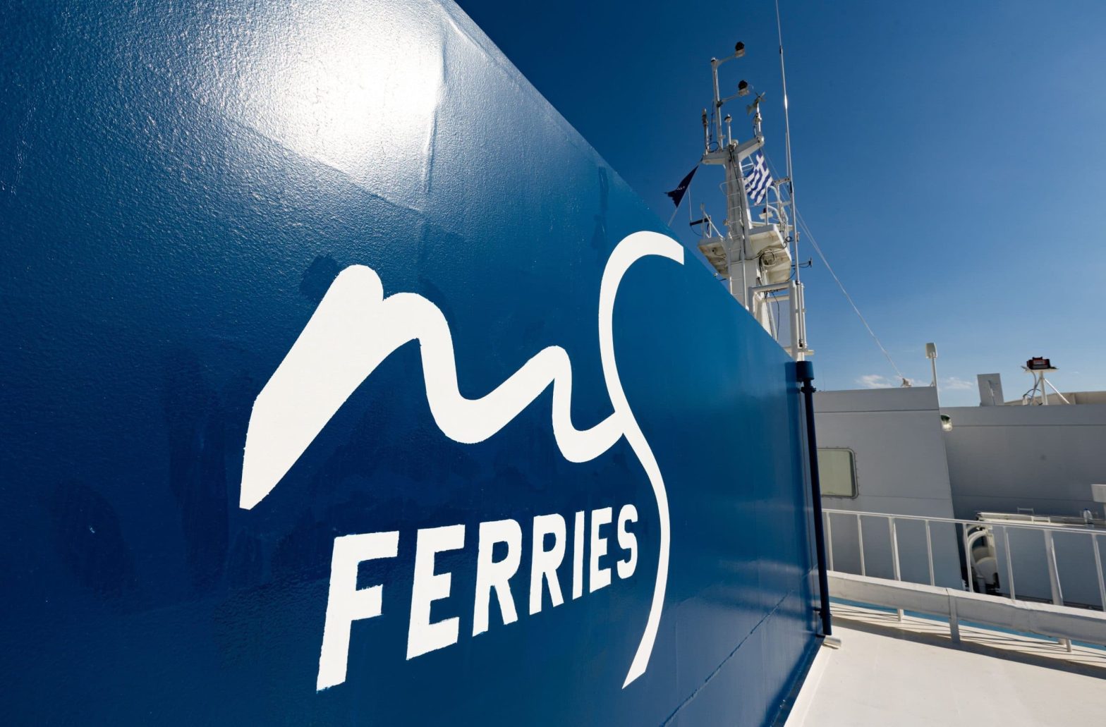 Close-up of the Magic Sea Ferries logo on a deep blue ferry exterior, with a Greek flag waving in the background under a clear blue sky.
