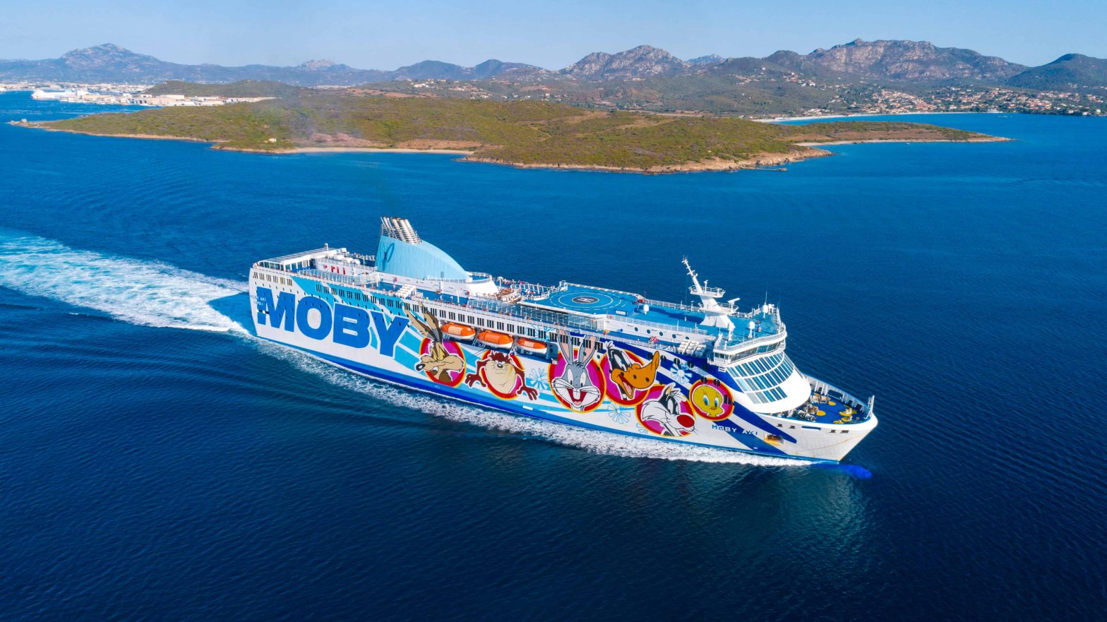 Traghetto Moby Aki in navigazione nelle acque azzurre del Mediterraneo, decorato con personaggi dei Looney Tunes e con un'isola verde sullo sfondo.