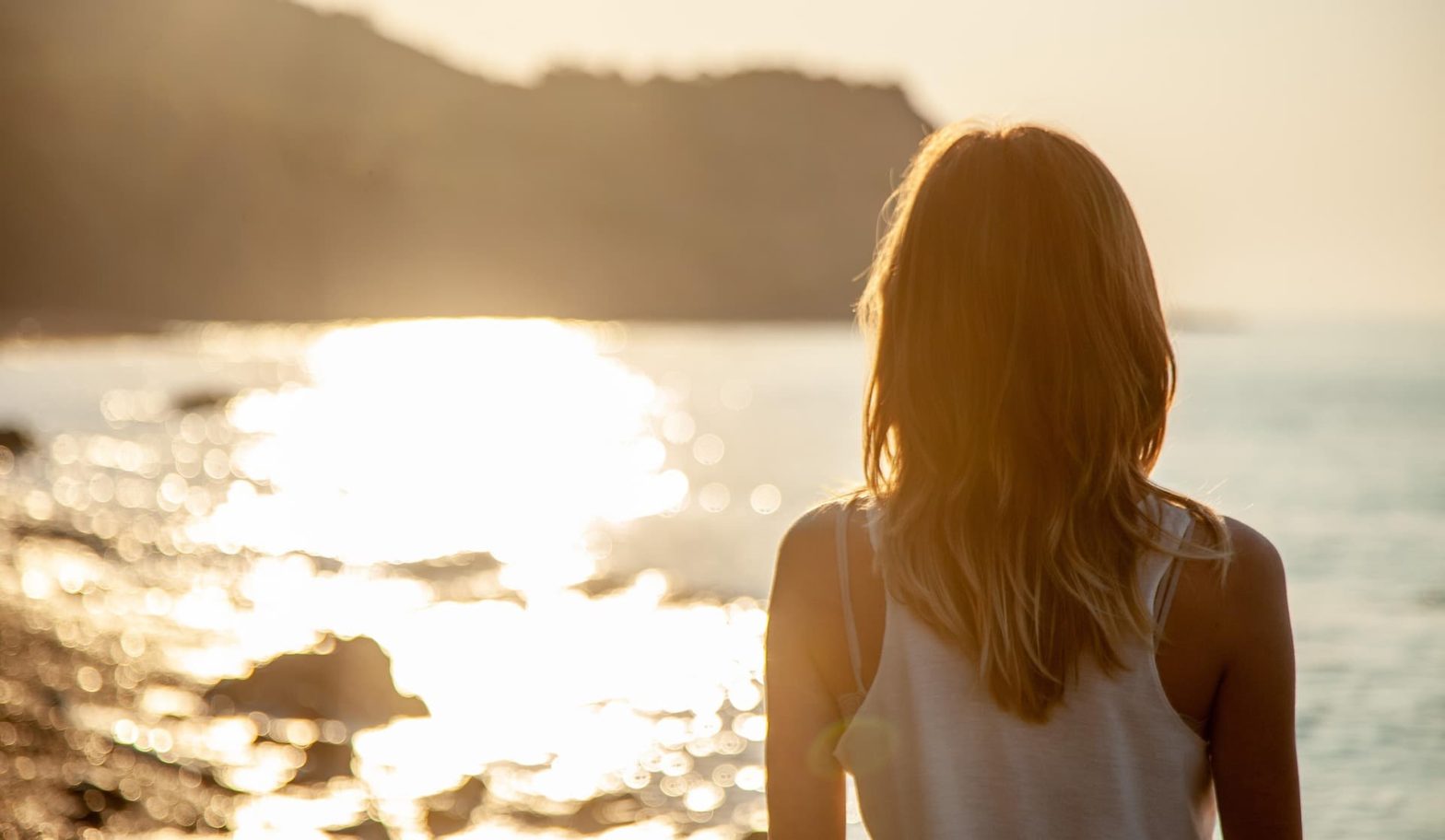 Donna di spalle in riva al mare al tramonto, mentre osserva l'acqua scintillante e la costa in lontananza.