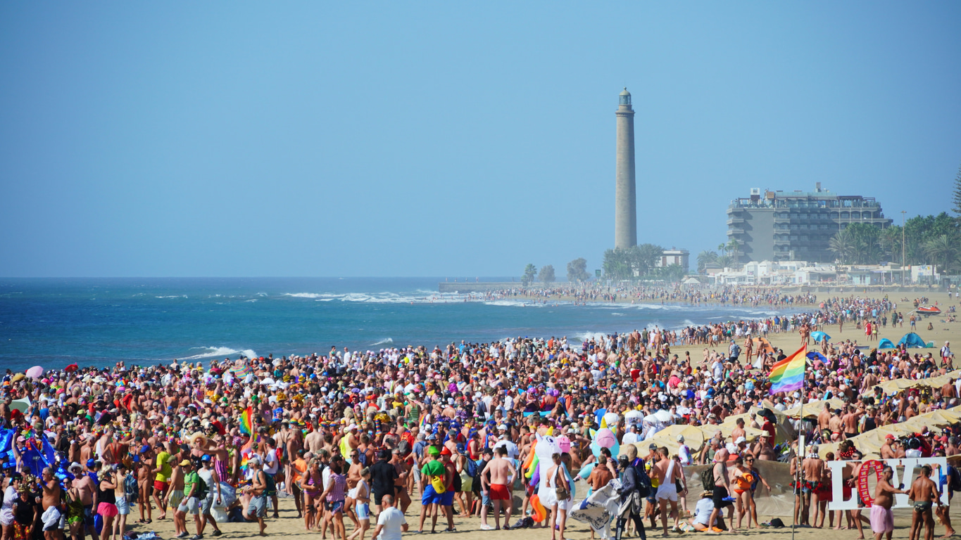Une foule aux couleurs LBGTQ+ sur la plage de Maspalomas avec le phare emblématique en fond.