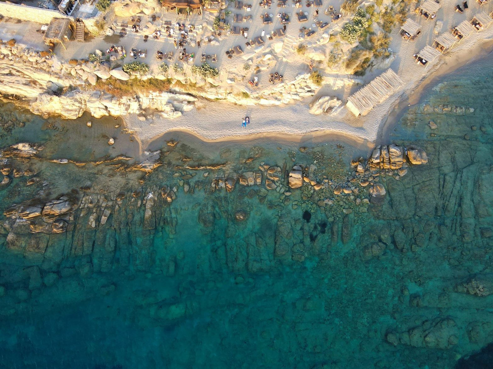 Vista aerea di una spiaggia di Mykonos con acqua cristallina, rocce sommerse e lettini disposti sulla sabbia.
