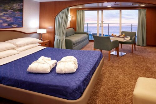 Cabina esterna di lusso su un traghetto con letto matrimoniale, area salotto, tavolo apparecchiato e ampie finestre vista mare al tramonto.