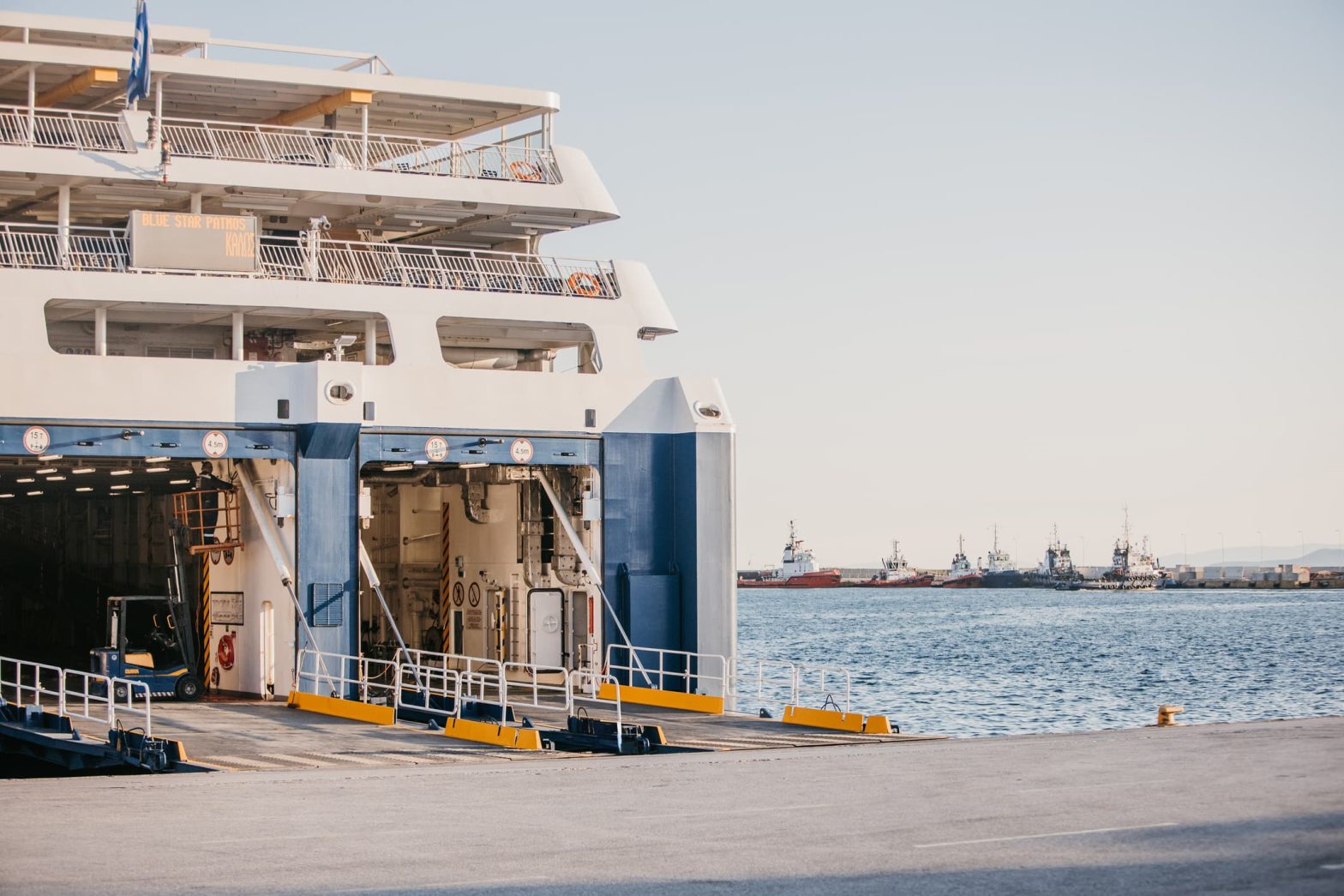 Traghetto attraccato al porto con rampa aperta per l’imbarco di veicoli, sullo sfondo il mare e diverse imbarcazioni ormeggiate
