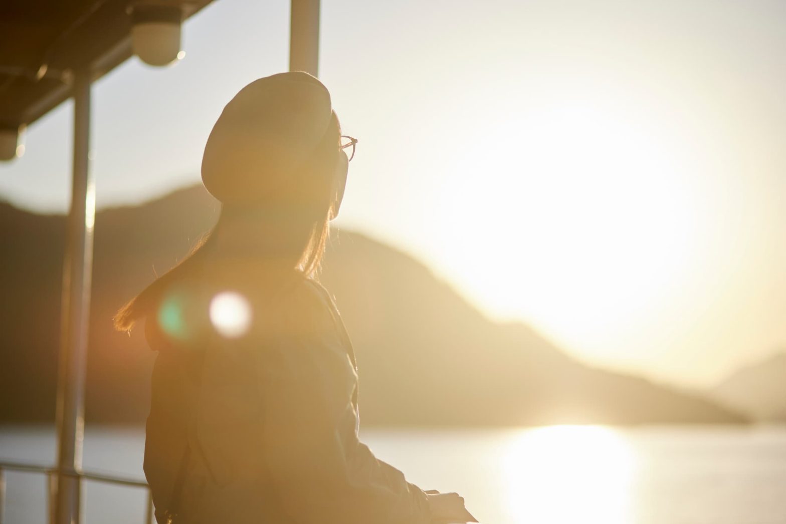 Donna con cappello e giacca osserva il mare al tramonto dal ponte di un traghetto.