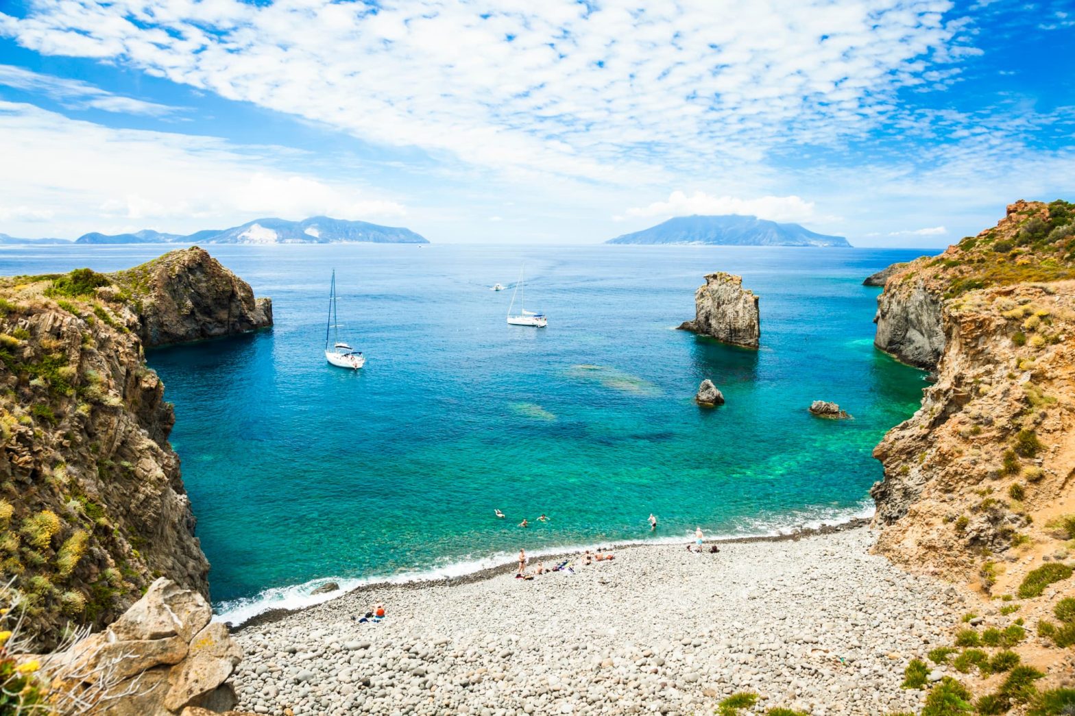 Vista panoramica su una spiaggia rocciosa con mare turchese e barche a vela ancorate tra le scogliere delle isole Eolie, con le isole vulcaniche sullo sfondo.