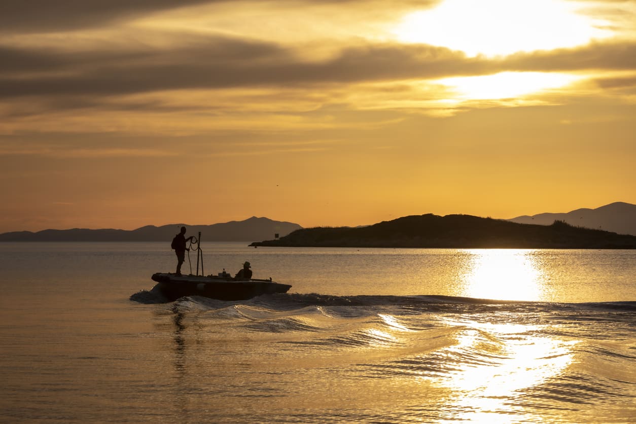 Zalazak sunca na moru koje okružuje otok Mljet sa jednom osobom koja vozi čamac i drugom koja stoji u čamcu koji za sobom ostavlja valove.